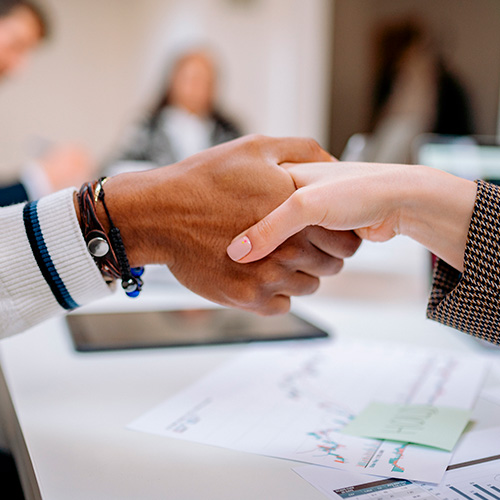 Duas pessoas apertando as mãos sobre uma mesa com documentos e gráficos, simbolizando acordo ou parceria profissional em ambiente corporativo.
