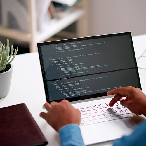 Pessoa digitando em notebook com tela exibindo linhas de programação, em ambiente de trabalho organizado com caderno e planta ao lado.
