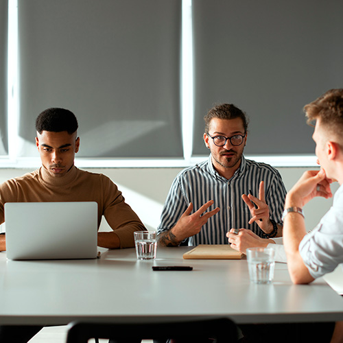 Grupo em reunião de trabalho, com pessoas conversando e usando notebook em mesa com copos de água.