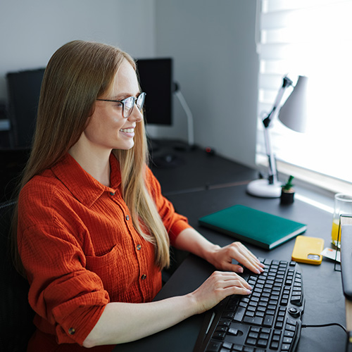 Mulher sorridente usa computador em mesa de trabalho, com objetos de escritório e luz natural entrando pela janela.