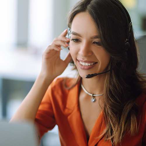 Atendente com headset sorri enquanto fala ao computador, realizando atendimento ao cliente em ambiente de trabalho.