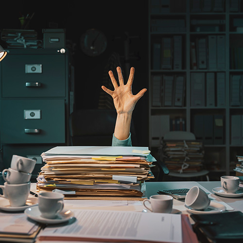 Mesa de escritório com pilhas de papéis e mão levantada ao fundo, representando sobrecarga e desorganização no trabalho.