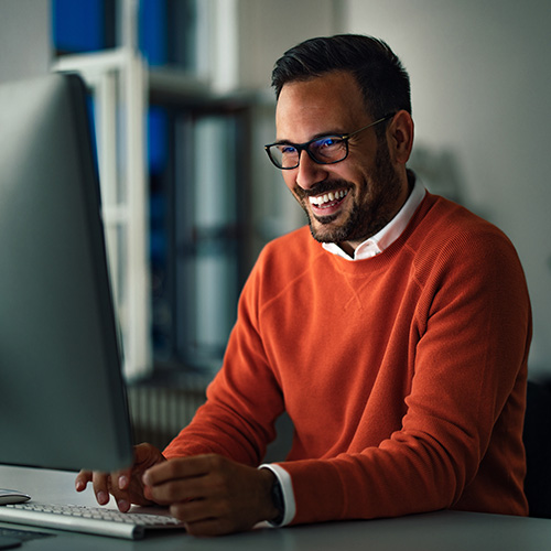 Homem sorridente usa computador em escritório, sentado à mesa com expressão satisfeita durante o trabalho.