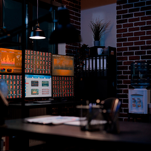 Escritório com mesa de trabalho, vários monitores exibindo gráficos e painéis de dados, estante com pastas, dispenser de água e iluminação em ambiente interno.